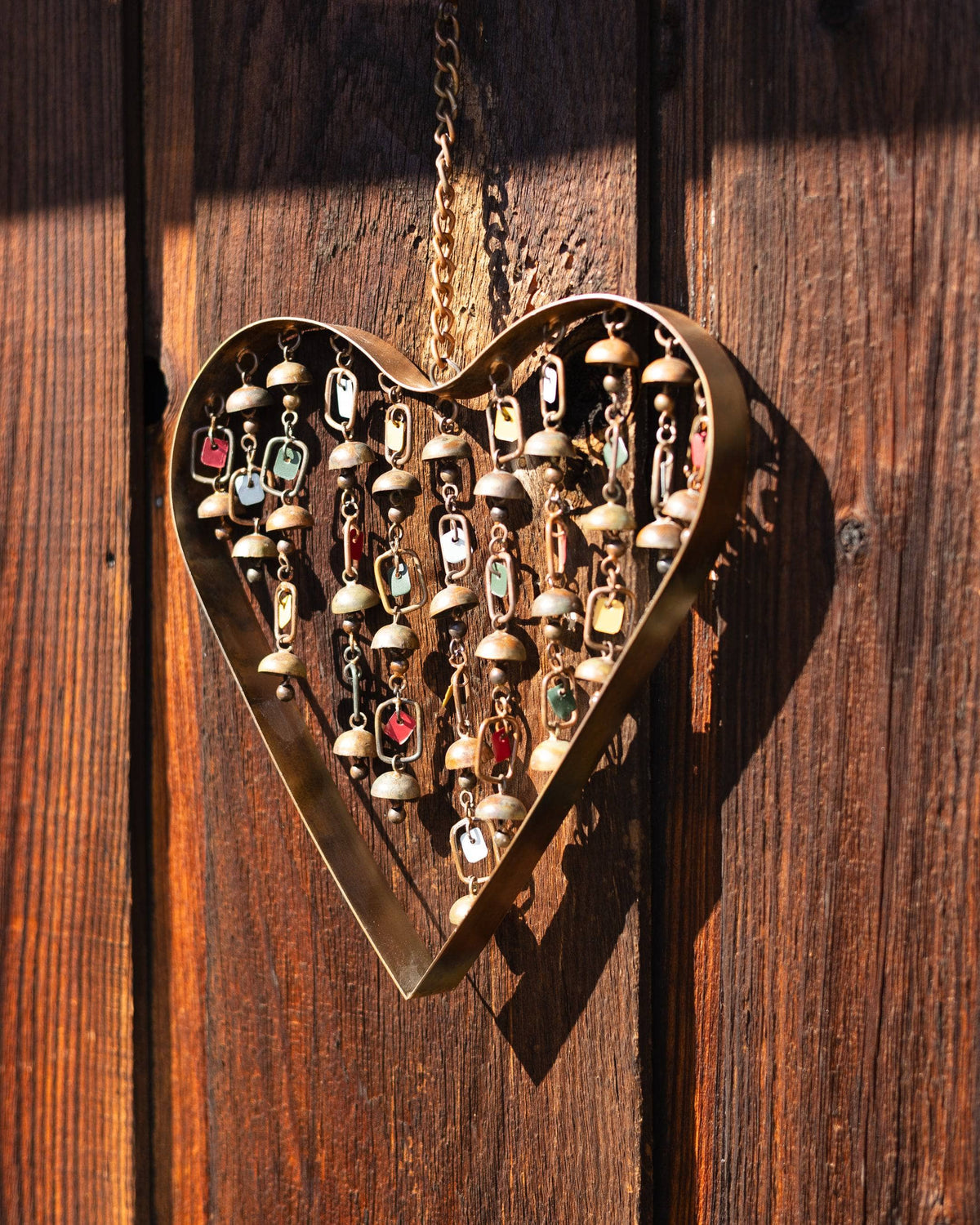 Gold metal heart-shaped wind chime with colorful dangling bells and charms, hung on a rustic wood fence