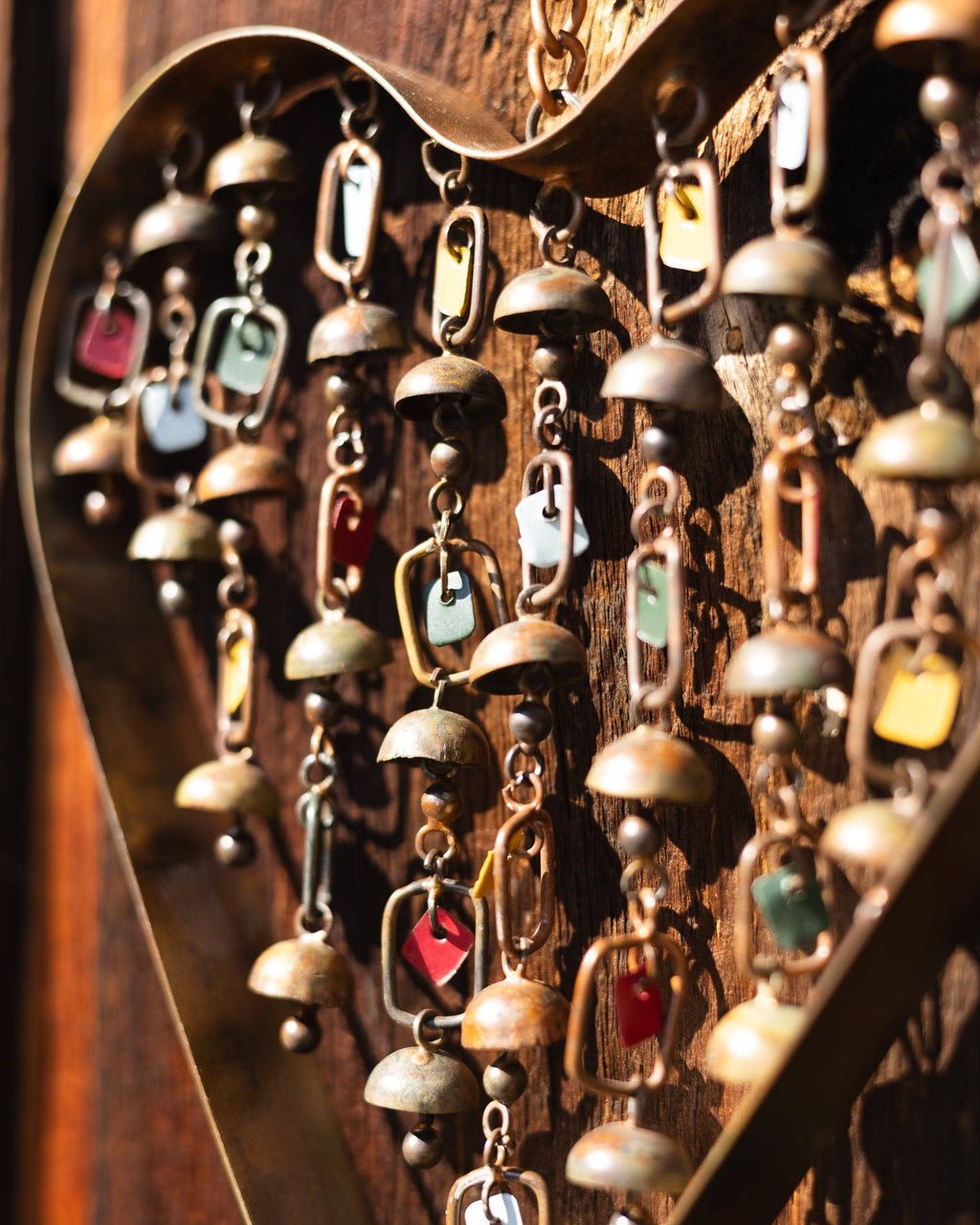 Metal heart-shaped wind chime with hanging brass bells and colorful enamel charms, suspended outdoors in autumn setting