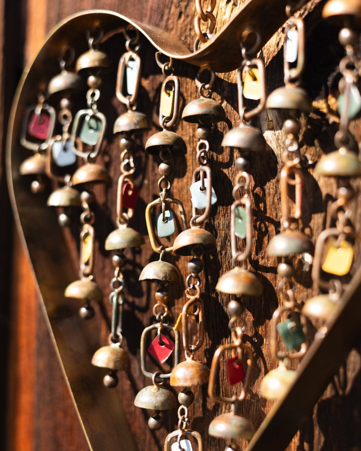 Metal heart-shaped wind chime with hanging brass bells and colorful enamel charms, suspended outdoors in autumn setting