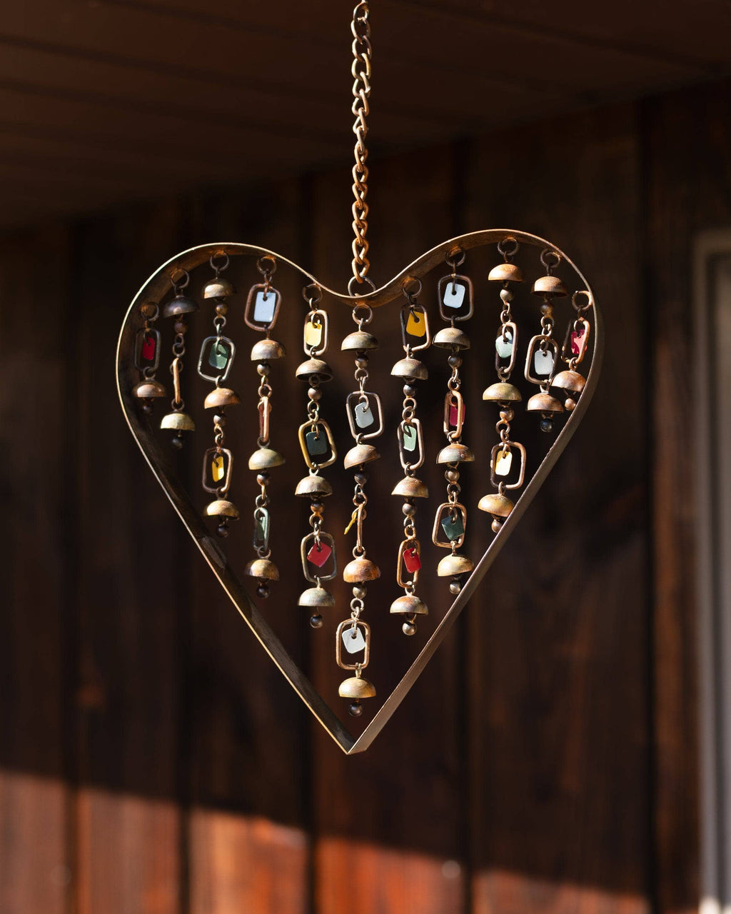 Brass heart-shaped wind chime with hanging bells and colorful tag dangles mounted on a wooden surface