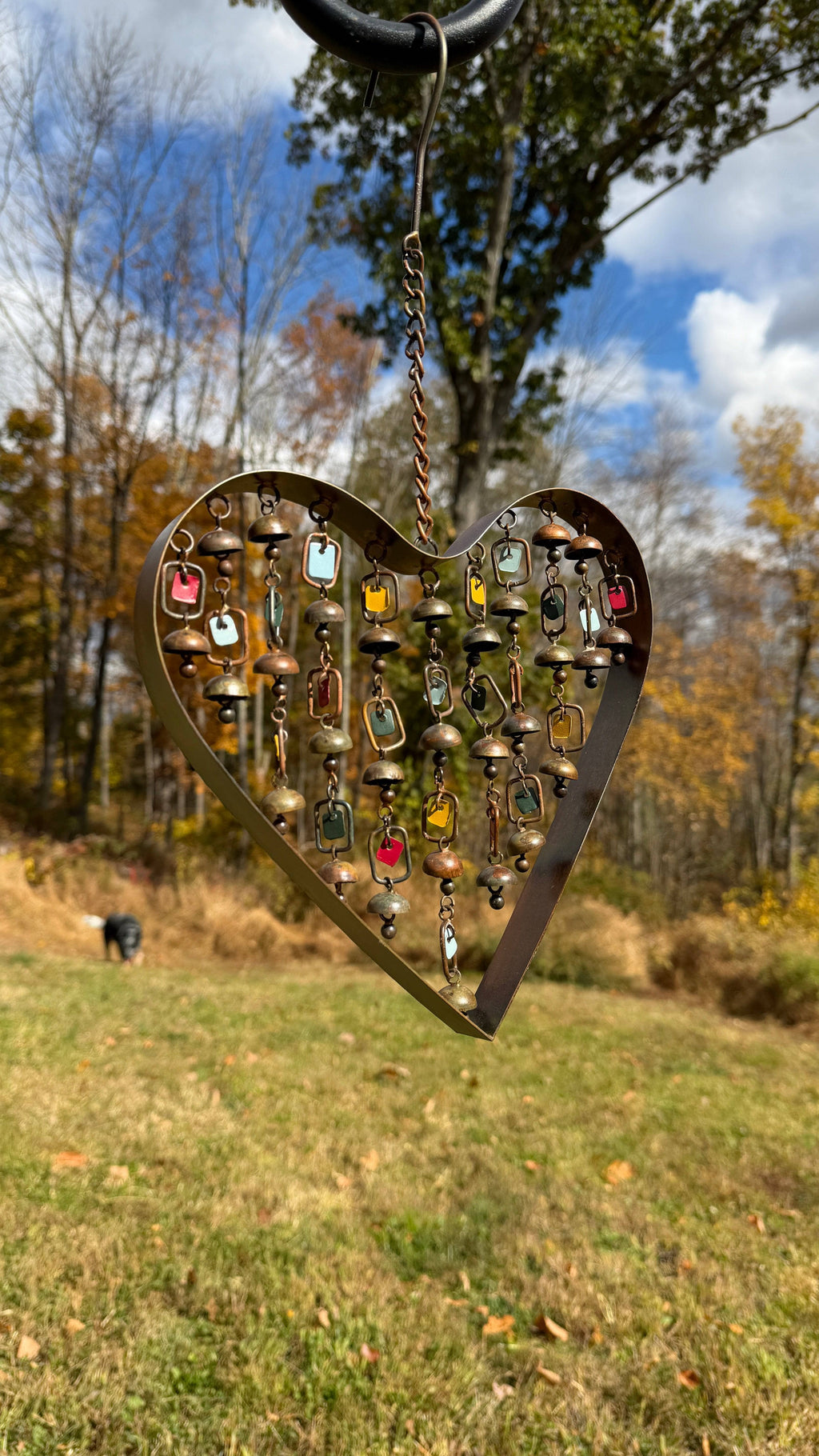 Brass heart-shaped wind chime with hanging small bells and colorful enamel tags, suspended by a chain against a wood wall
