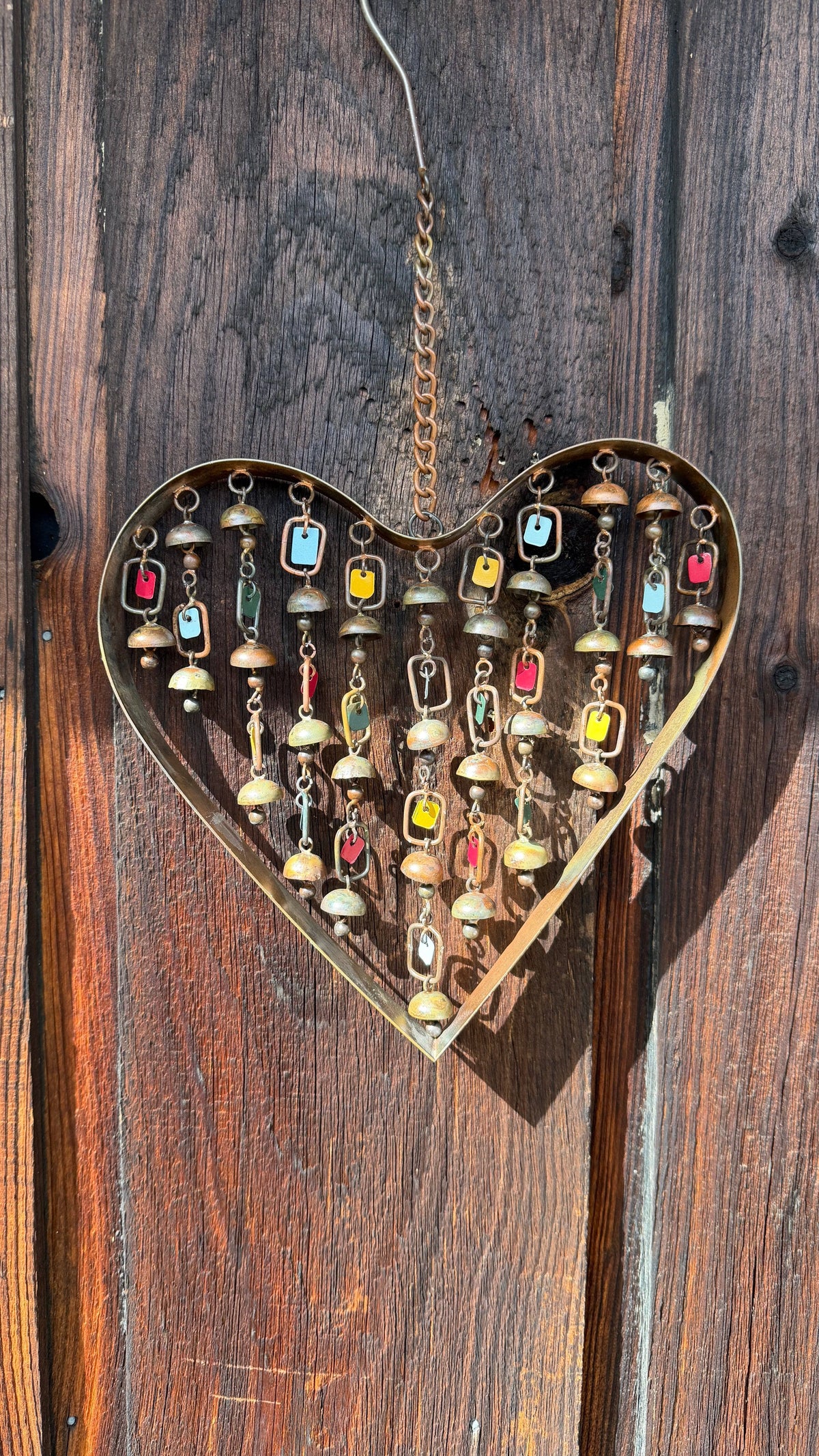 Brass heart-shaped wind chime with hanging chains, small bells, and colorful enamel tags, suspended from a metal chain