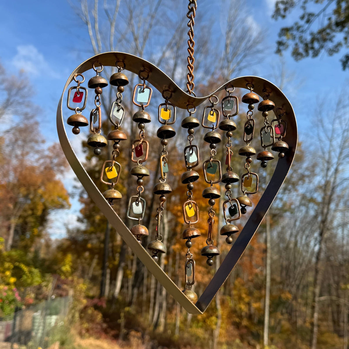 Copper heart-shaped wind chime with hanging chains, small bells, and colorful enamel tags, suspended outdoors